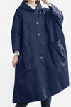 A person is wearing the Navy Blue One Size Fits All Oversized Waterproof Windbreaker Jacket by looselyboho. The jacket is long and loose-fitting with large front pockets and a hood. The oversized fit adds to its stylish look, featuring large buttons down the front. The person's hands are partially visible from the sleeves, with one hand holding the hood. The background is plain white.