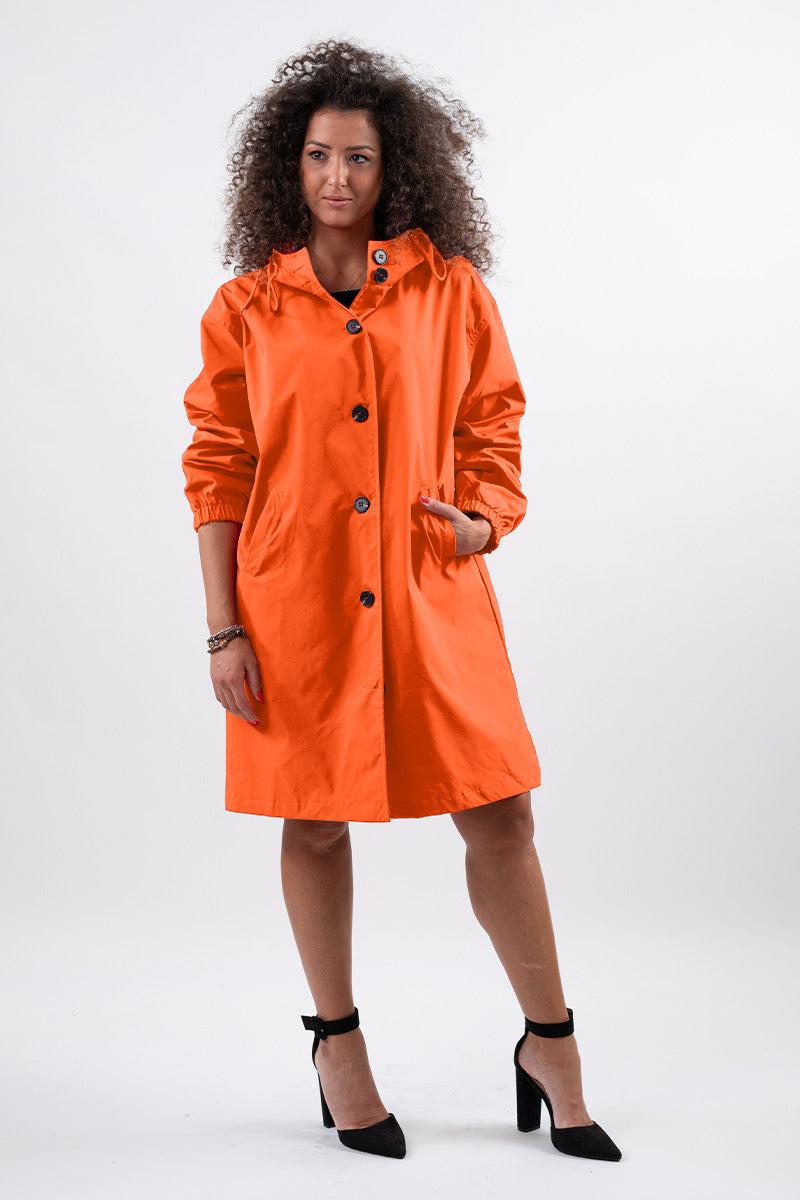 A person with curly hair is dressed in a PUMPKIN ORANGE WINDBREAKER by looselyboho and black heels. They are positioned against a plain white background, with one hand casually tucked into a pocket and the other resting at their side.